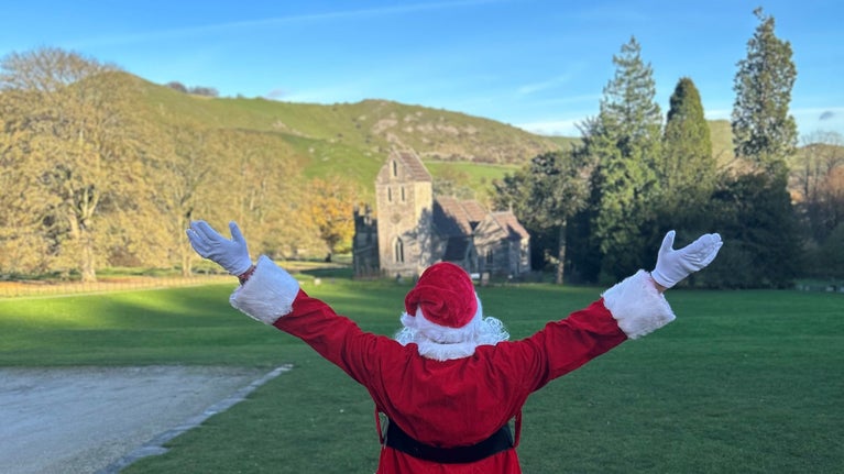 The back of Santa in his red suit with his arms in the air and open wide as he looks at the view of hills and a church in the distance